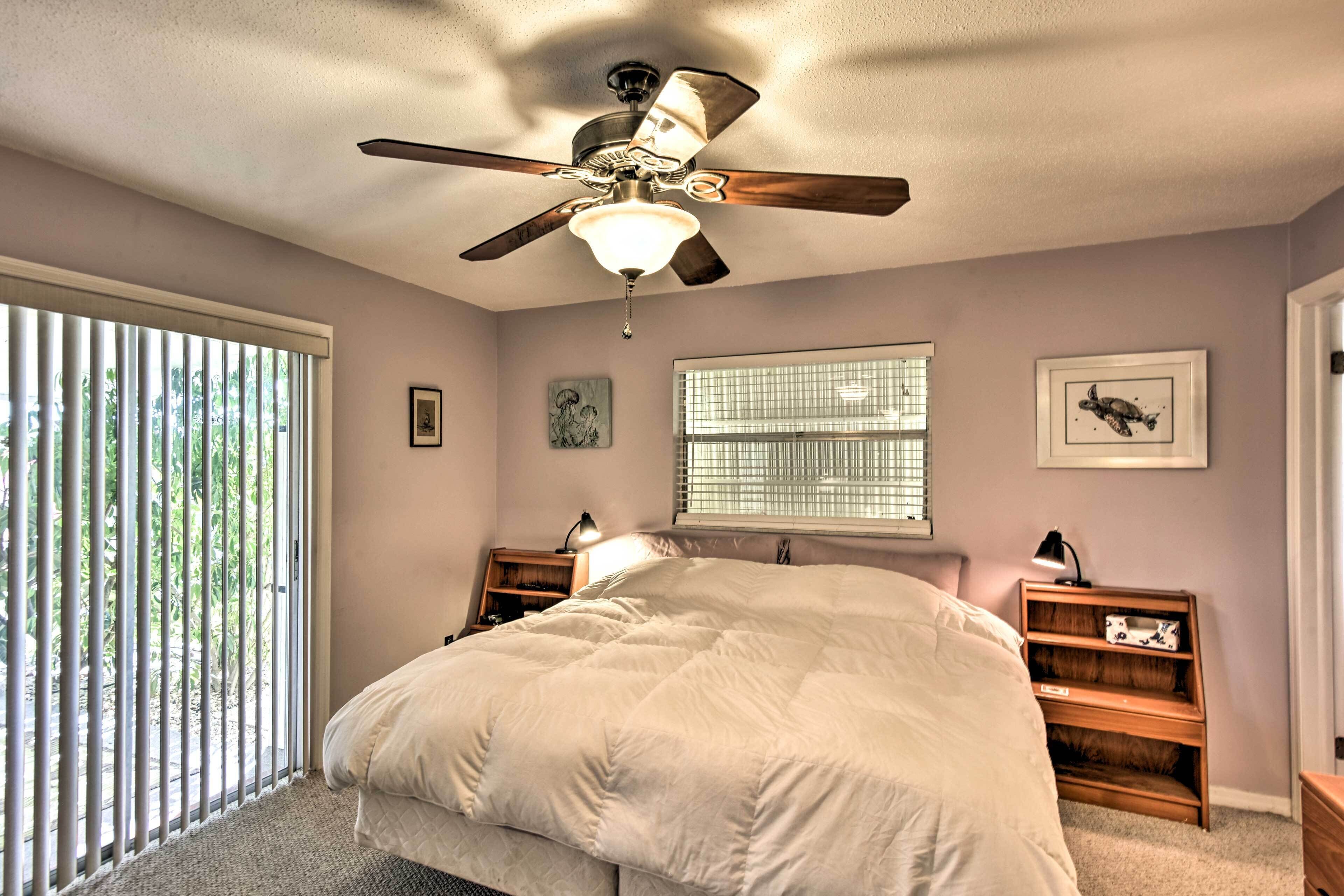 A bedroom with a bed, ceiling fan, window with blinds, bedside tables, and wall art.