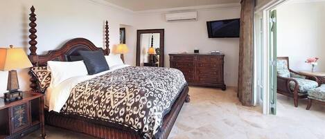 Un dormitorio amplio con una cama grande, cabecera de madera, ventilador de techo, televisor, cómoda y una silla.
