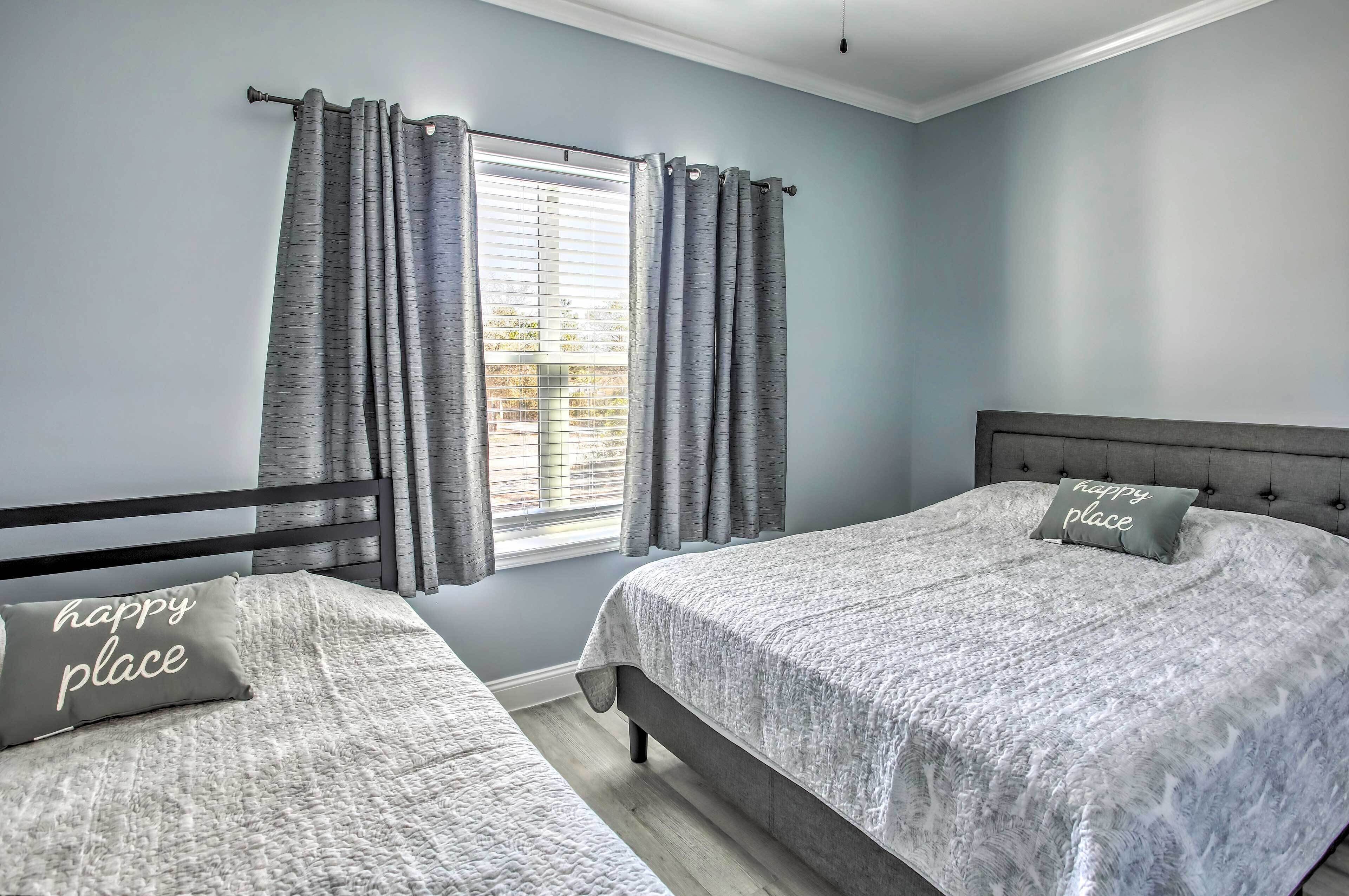 A bedroom with two beds, grey curtains, a window with blinds, and a pillow with the text "happy place".