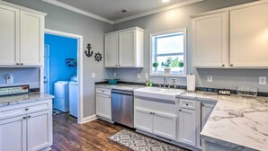 A modern kitchen with white cabinets, a marble countertop, a built-in dishwasher, and a window with a view of greenery. - Ocean Isle Beach Escape - Centrally Located! (Ocean Isle Beach)