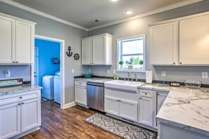 A modern kitchen with white cabinets, a marble countertop, a built-in dishwasher, and a window with a view of greenery. - Ocean Isle Beach Escape - Centrally Located! (Ocean Isle Beach)