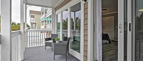 A covered balcony with a white railing, a seating area with wicker chairs, and a sliding glass door leading to a room with a bed and a nightstand.