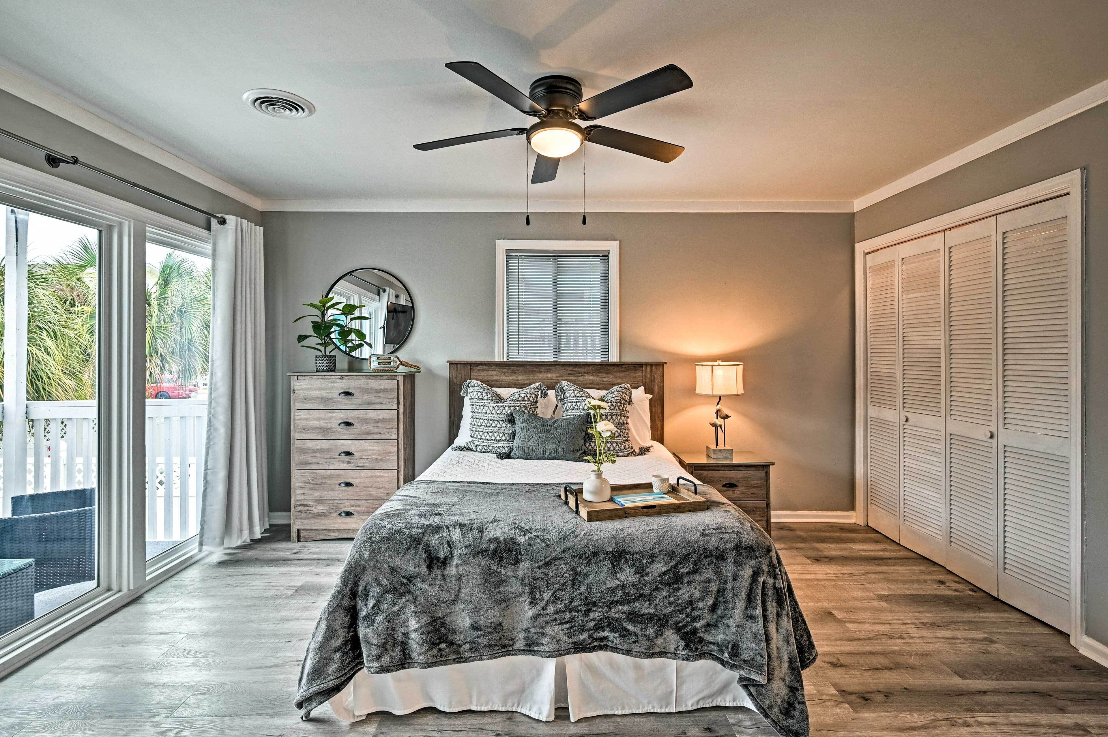 Quarto com cama, ventoinha no teto, mesa de cabeceira com abajur, cômoda e janela com cortinas.