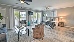Ein geräumiges Wohnzimmer mit einer Couch, einem Couchtisch und einem Korbstuhl. Ein Bett mit Nachttisch und Lampe ist ebenfalls vorhanden. Das Zimmer verfügt über große Fenster und einen Deckenventilator.