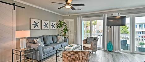 Un salon avec un canapé gris, une table basse, un fauteuil en osier, une lampe sur pied et un ventilateur de plafond. Des décorations en forme d’étoiles ornent le mur, et une grande fenêtre est agrémentée de rideaux.