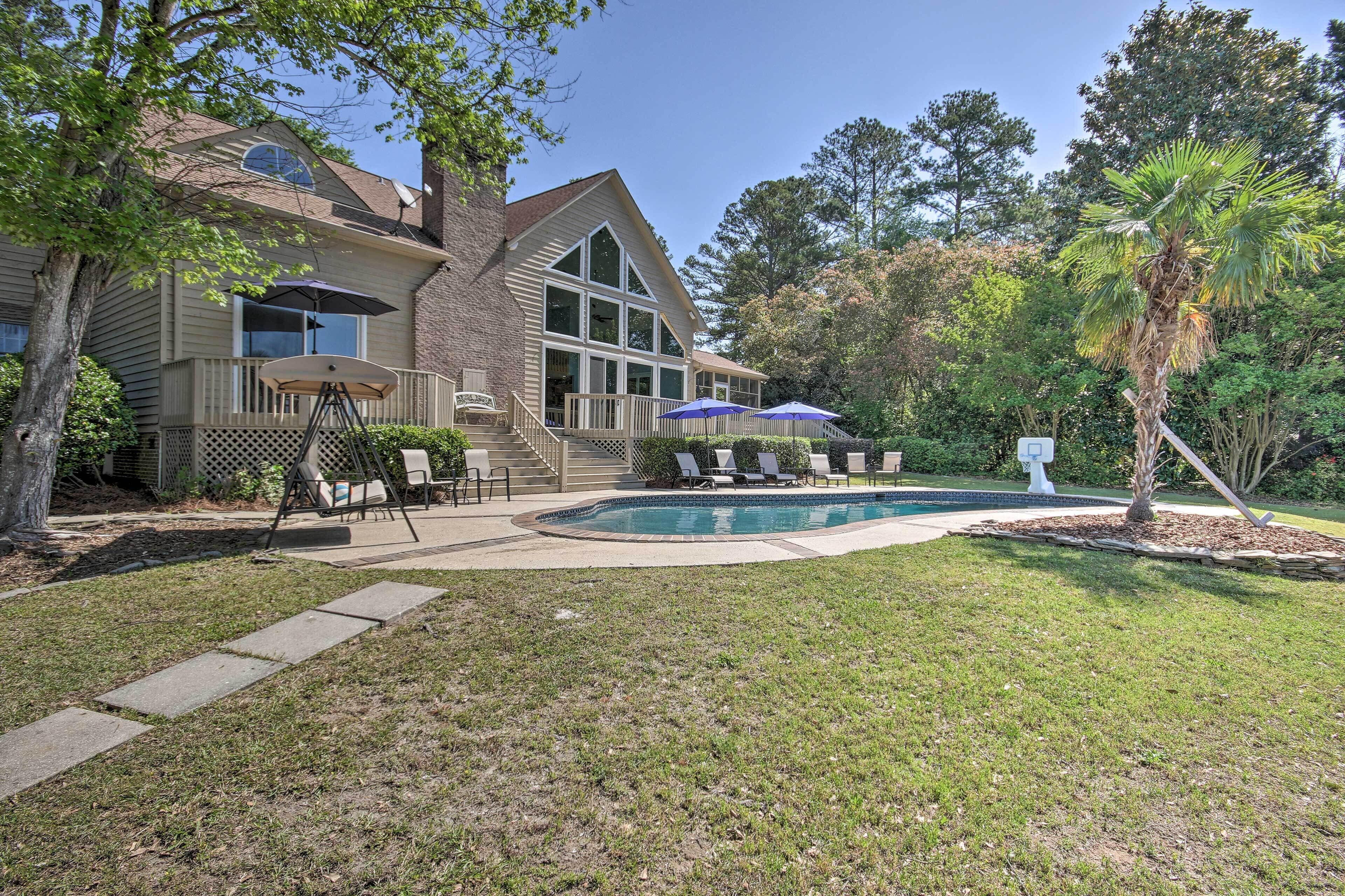 A house with a swimming pool, a covered patio with chairs and umbrellas, and a well-maintained lawn.