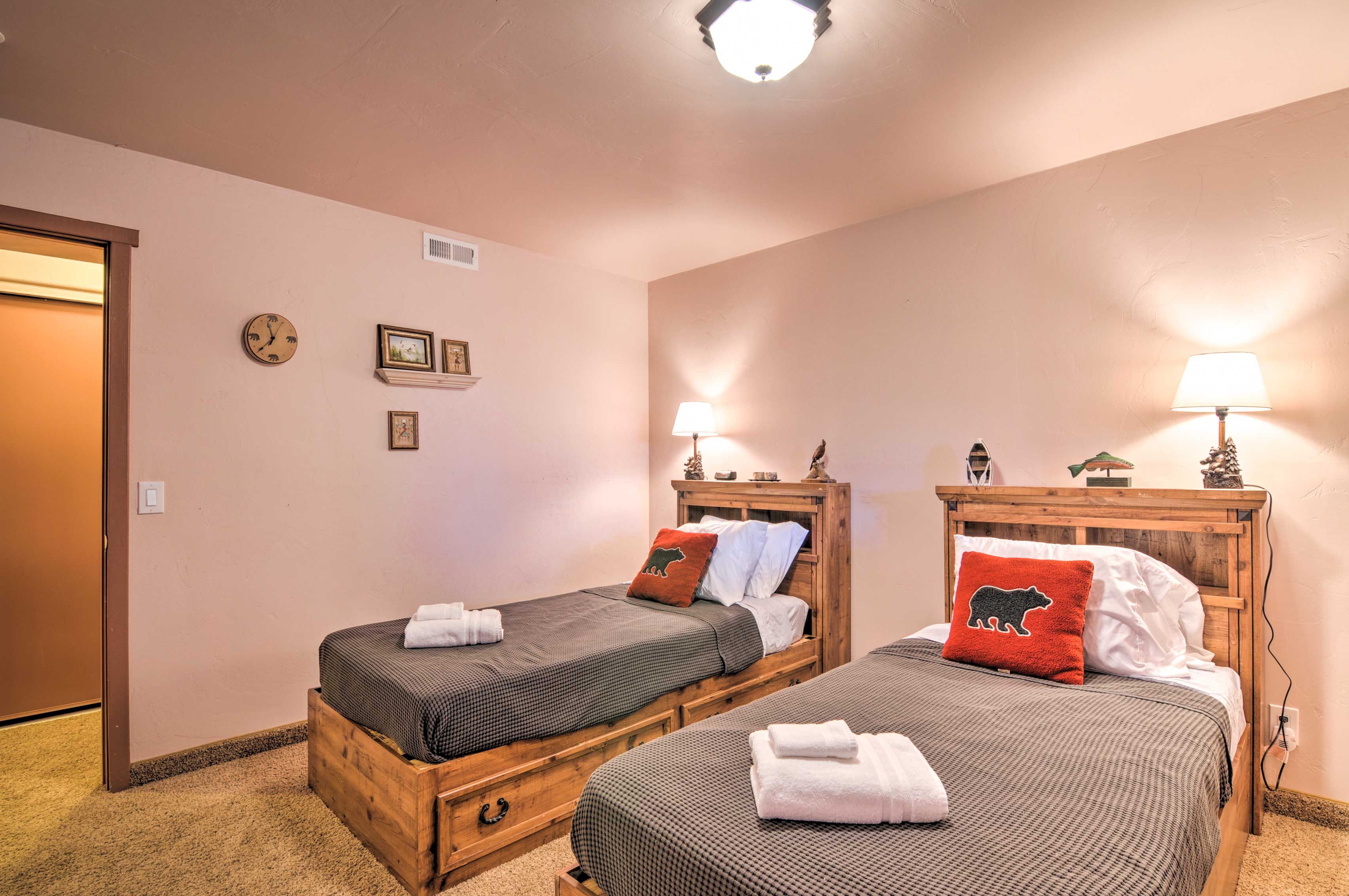 A hotel room with two wooden beds, each with a pillow and a lamp. There is a clock on the wall and a door to the left.