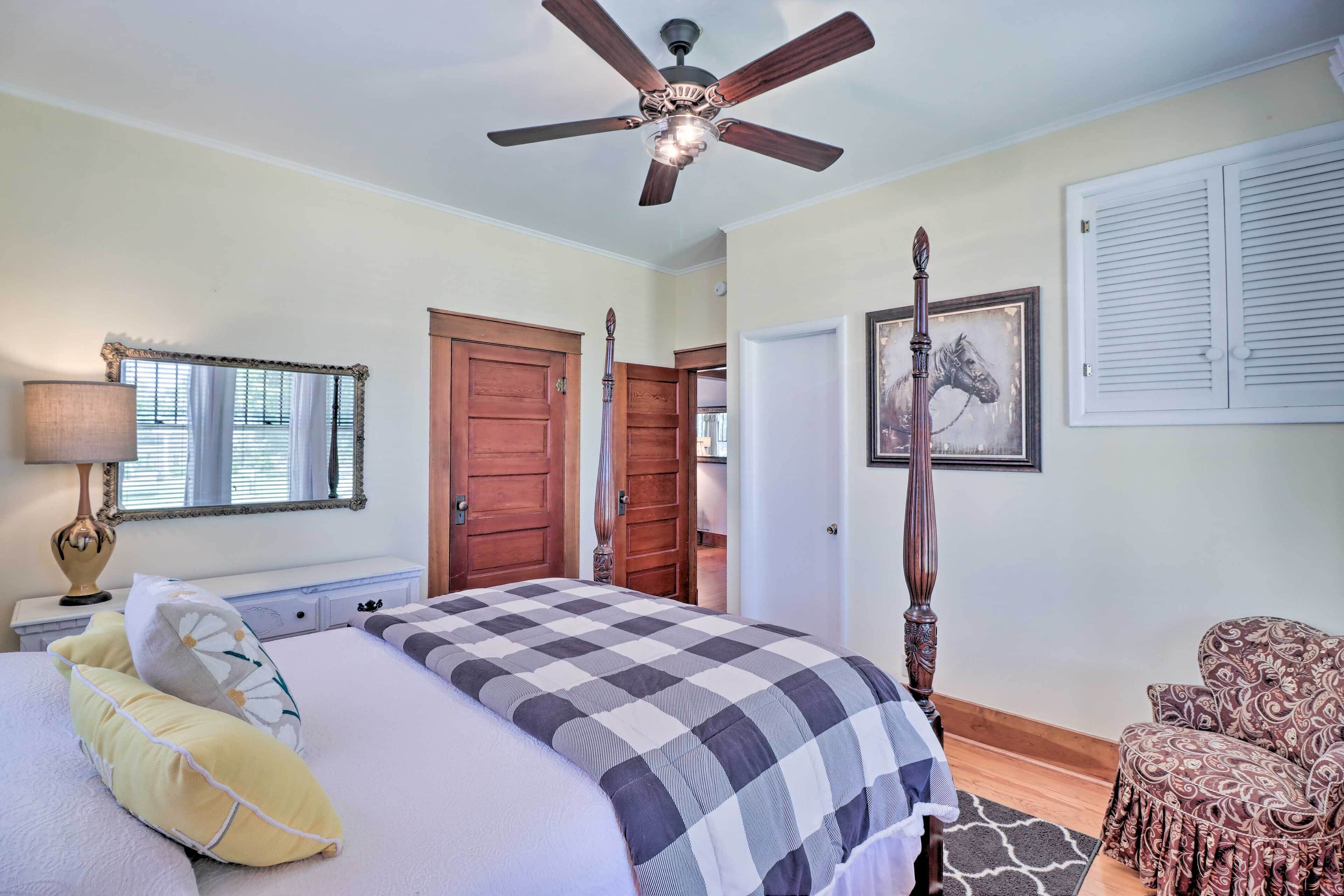 Une chambre à coucher avec un lit, un ventilateur de plafond, une porte en bois, un tableau encadré et une chaise à motifs.