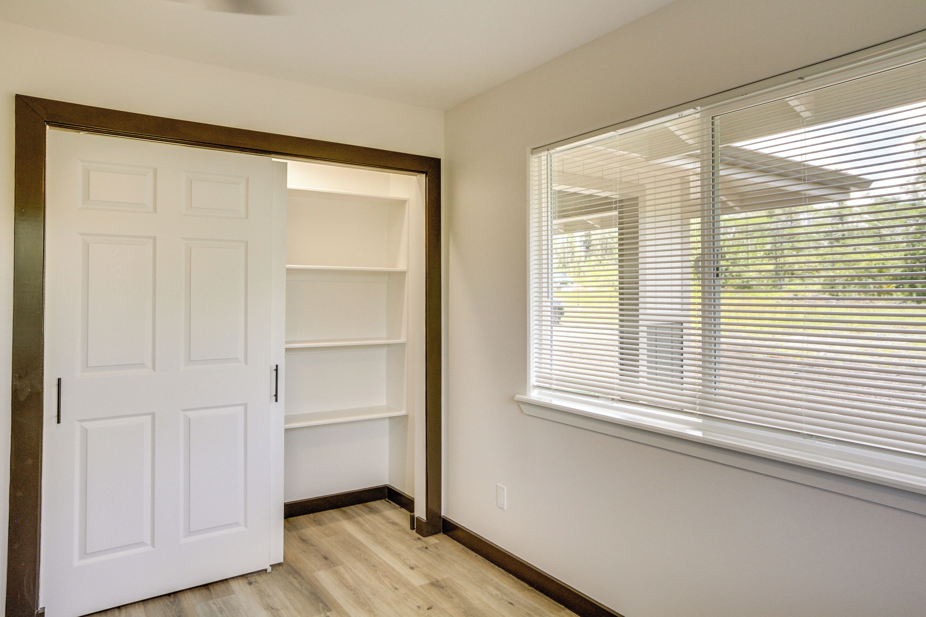 A room with a white door, a window with blinds, and a wooden floor.