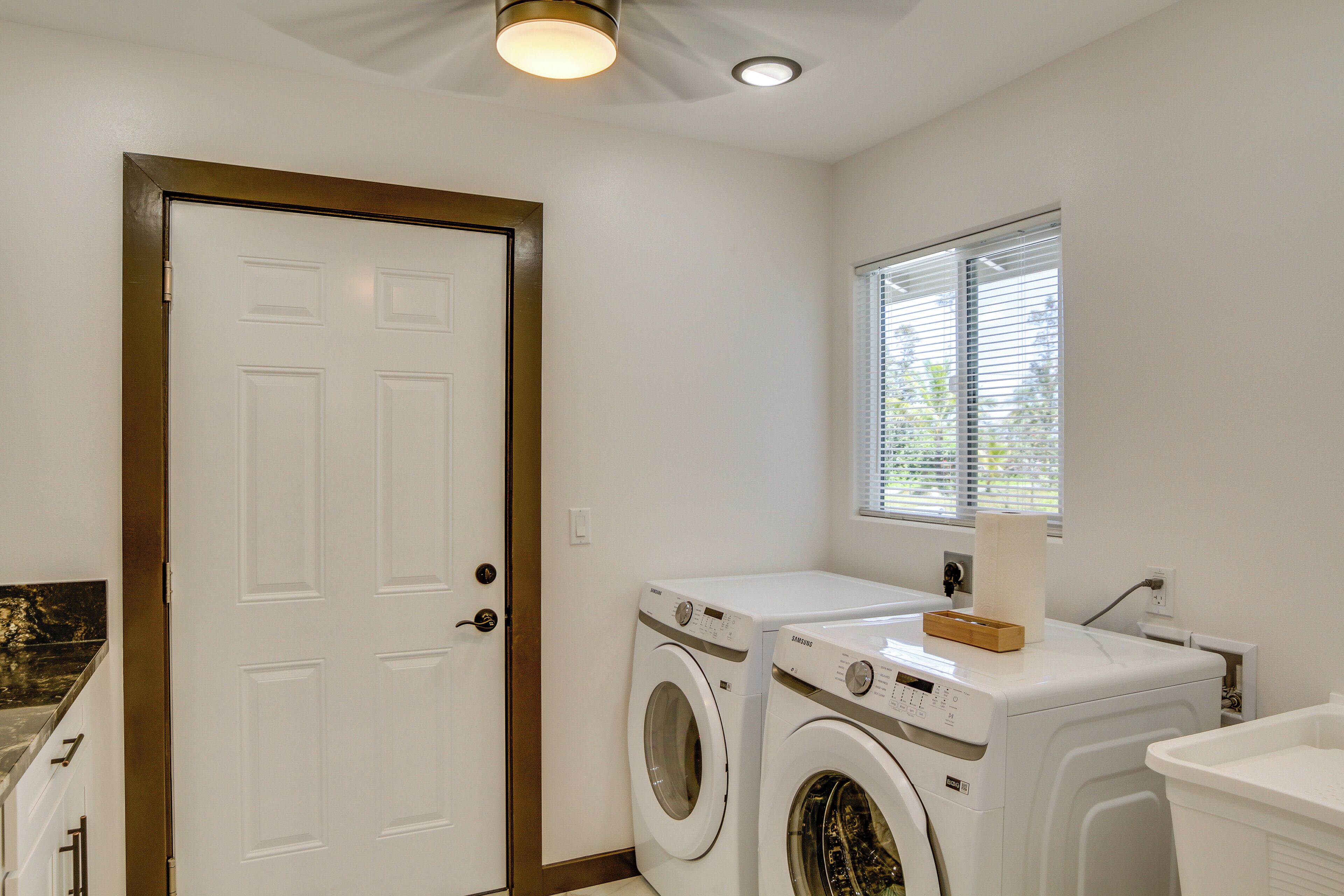 A laundry room with two front-loading washing machines, a white door, and a window with blinds.