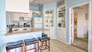 Una cocina con una isla central, taburetes de bar, muebles blancos y vistas al baño contiguo.