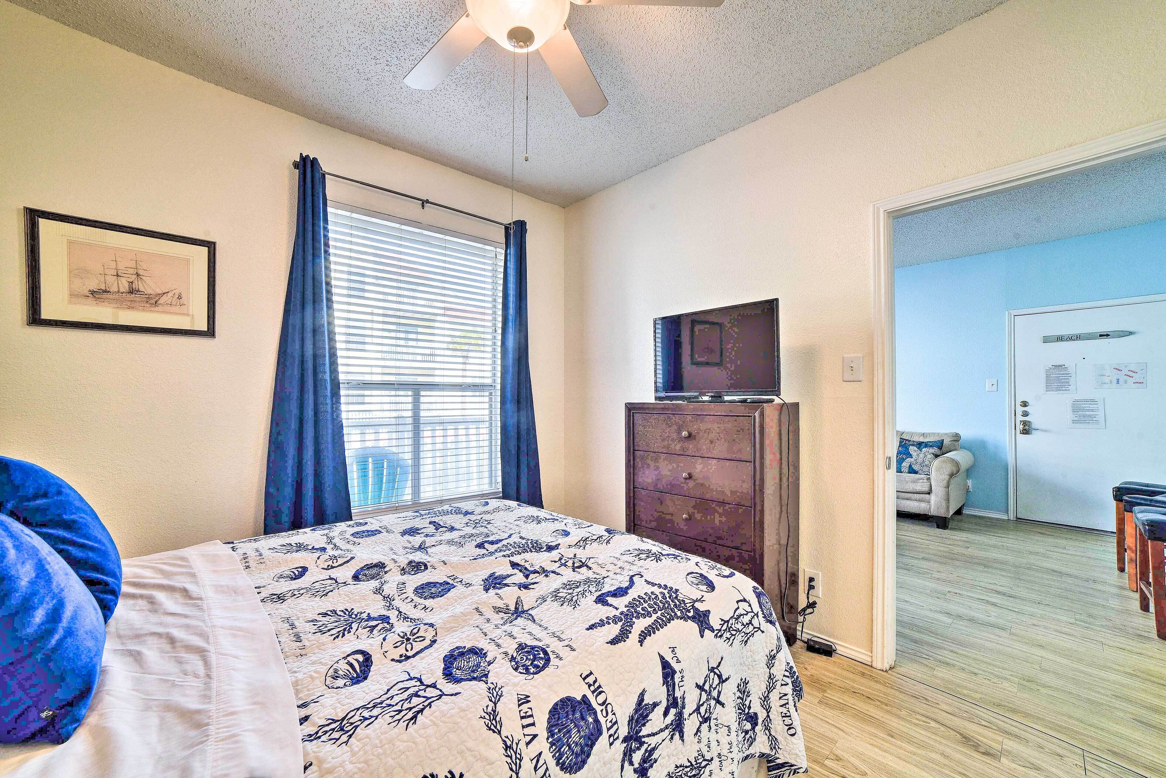 A bedroom with a bed, a dresser, a ceiling fan, a window with curtains, a framed picture on the wall, and a door leading to another room.