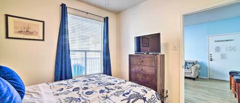 A bedroom with a bed, a dresser, a ceiling fan, a window with curtains, a framed picture on the wall, and a door leading to another room.