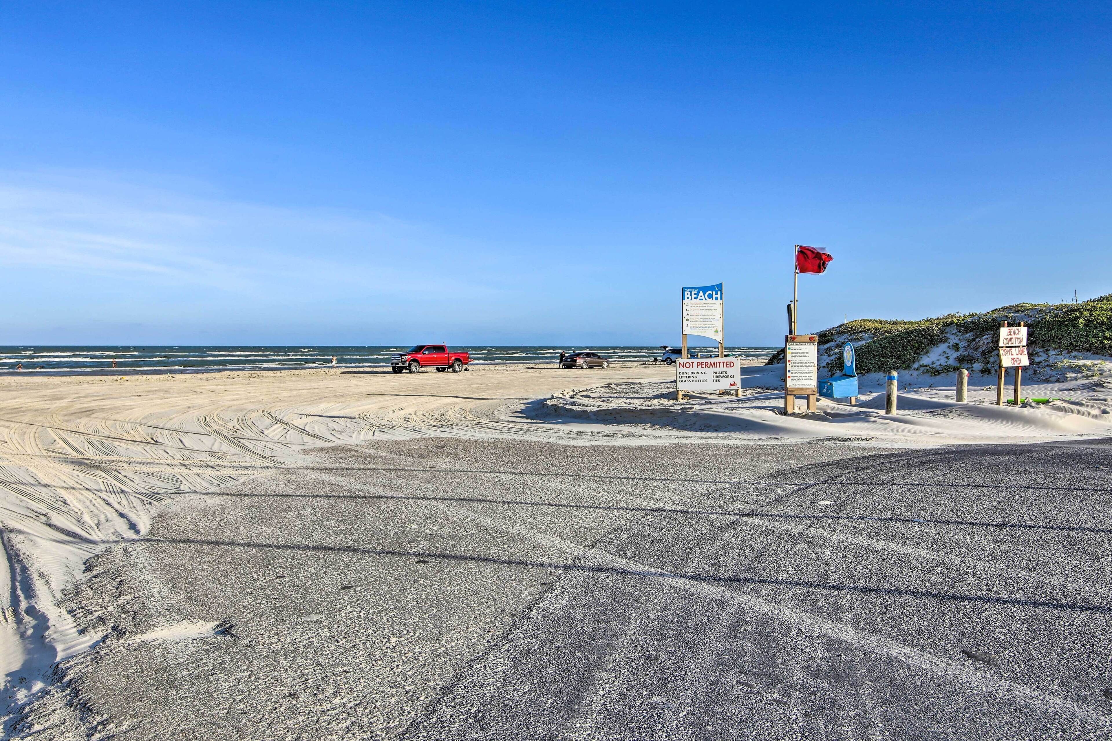 Una playa con un cartel que indica acceso a la playa, un coche rojo y una bandera.