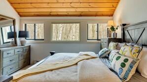 Une chambre avec un plafond en bois, un lit agrémenté d’oreillers à motifs, une table de chevet avec une lampe et des fenêtres donnant sur des arbres.