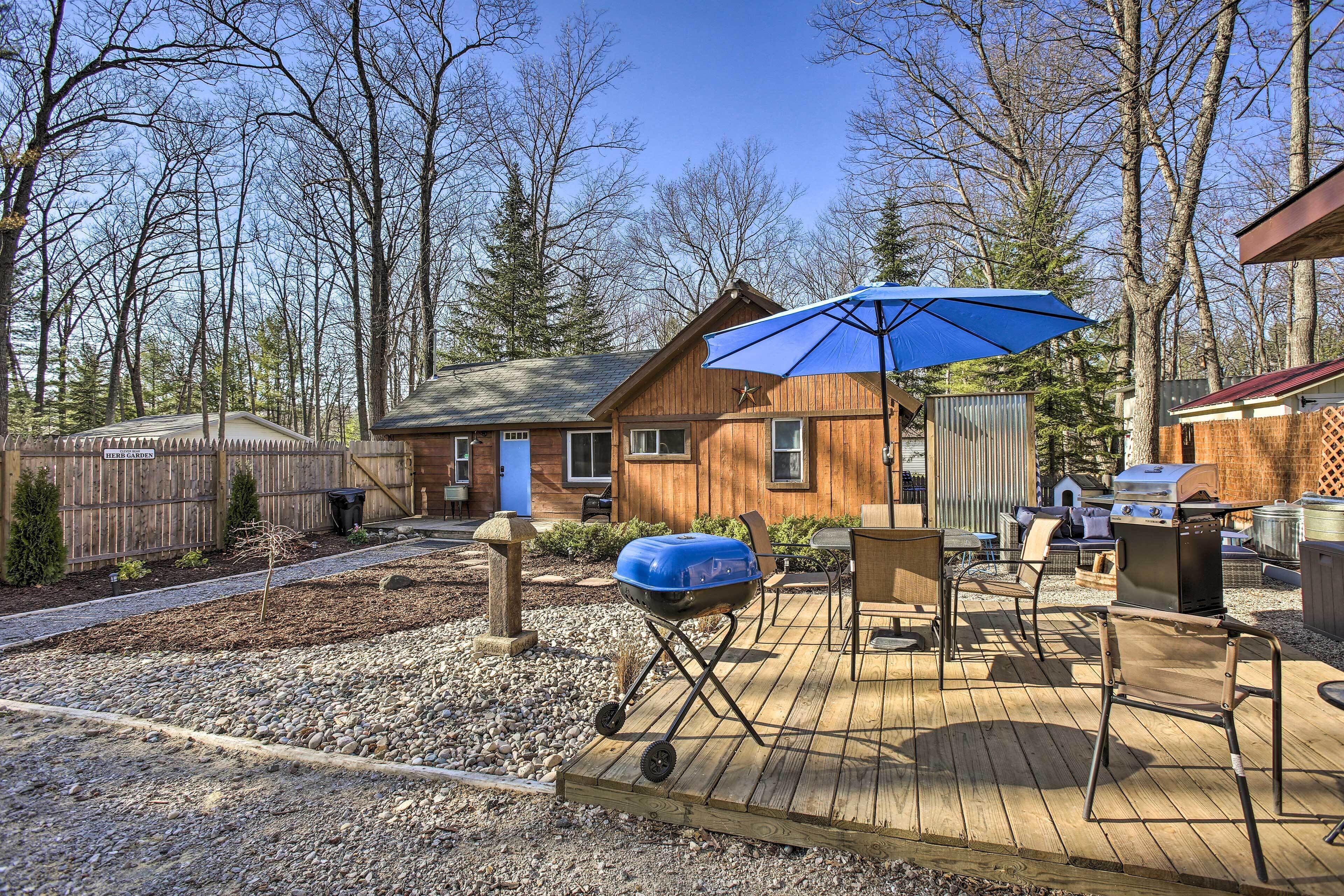 A wooden deck with a grill and barbecue, a blue umbrella, and a wooden house in a wooded area.