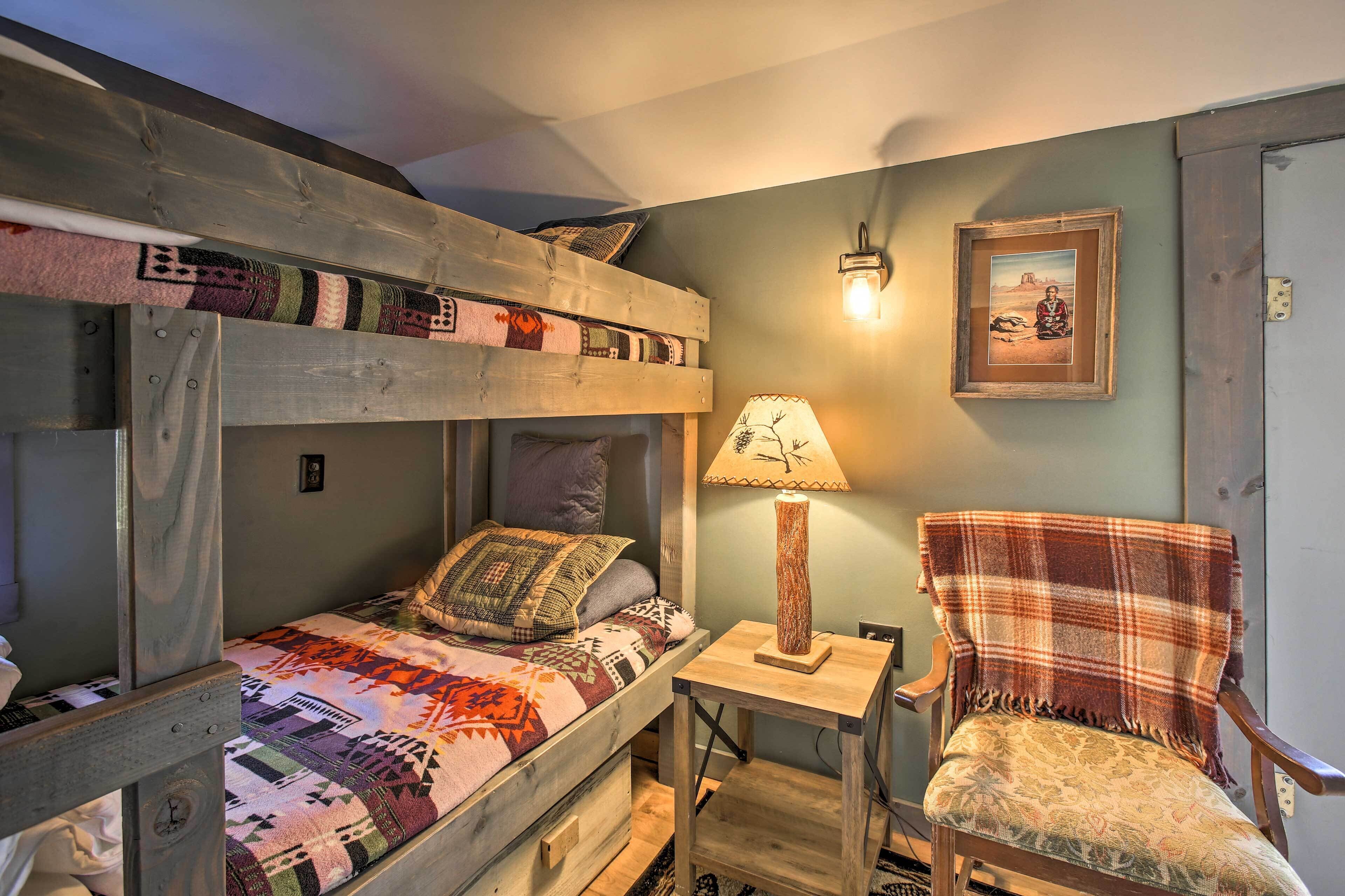 Een stapelbed met een geborduurd plaid, een houten nachtkastje met een lamp, en een stoel met een ruitdessin plaid.