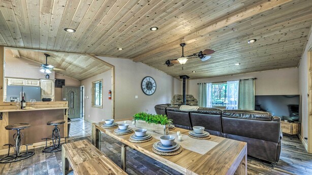 Amplia sala de estar con techo de madera, un sofá grande, una mesa de comedor con cuencos y platos, y una zona de cocina con un reloj y armarios.