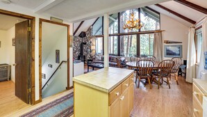 A spacious kitchen with a large island, wooden dining table, and chairs, a stone fireplace, and large windows with curtains.