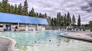 A swimming pool area with a blue-roofed building, lounge chairs, and a forested backdrop.