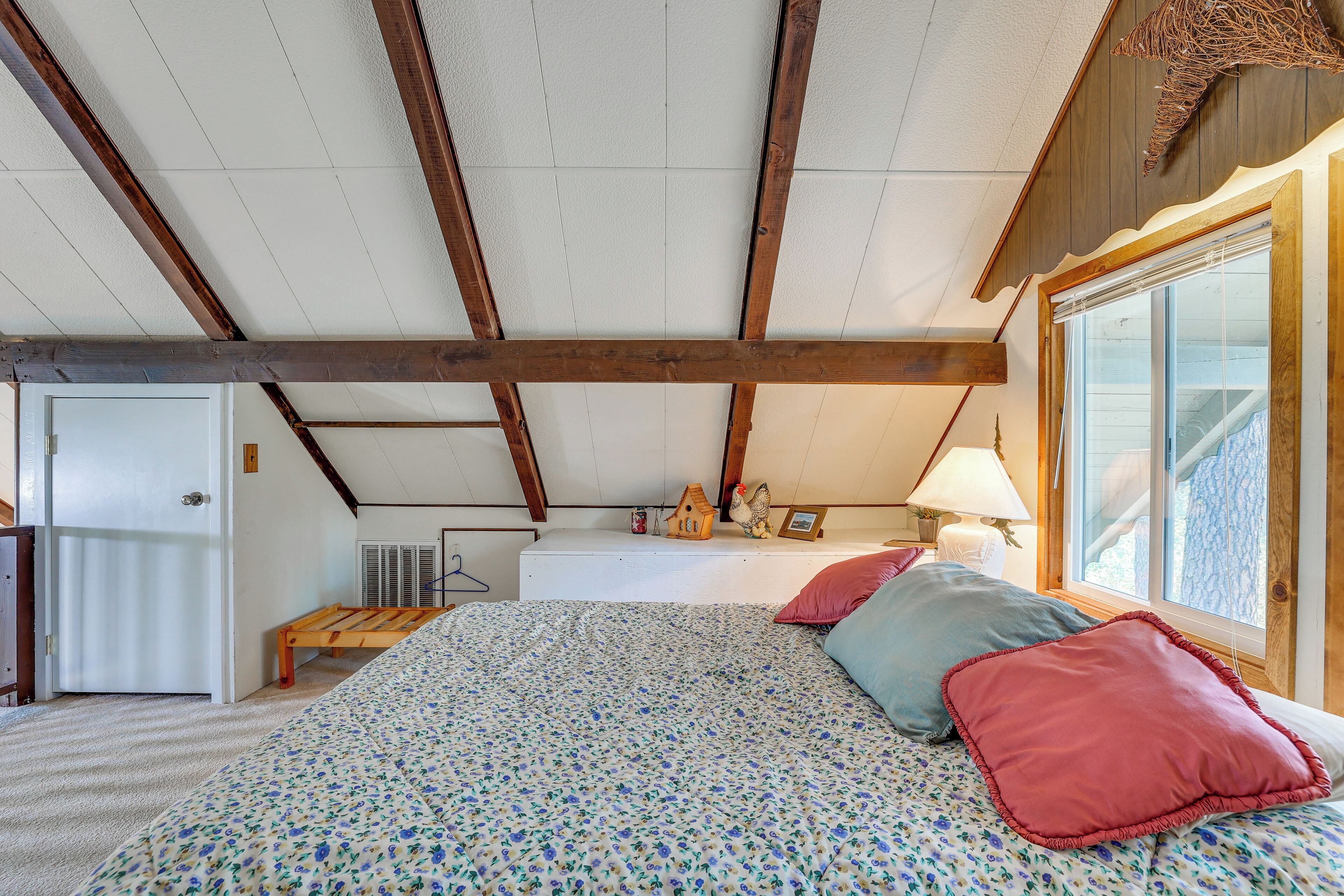 Une chambre avec un plafond en pente, un lit avec une literie à motifs floraux, des oreillers, une table de chevet, une lampe et une fenêtre donnant sur un beau paysage.