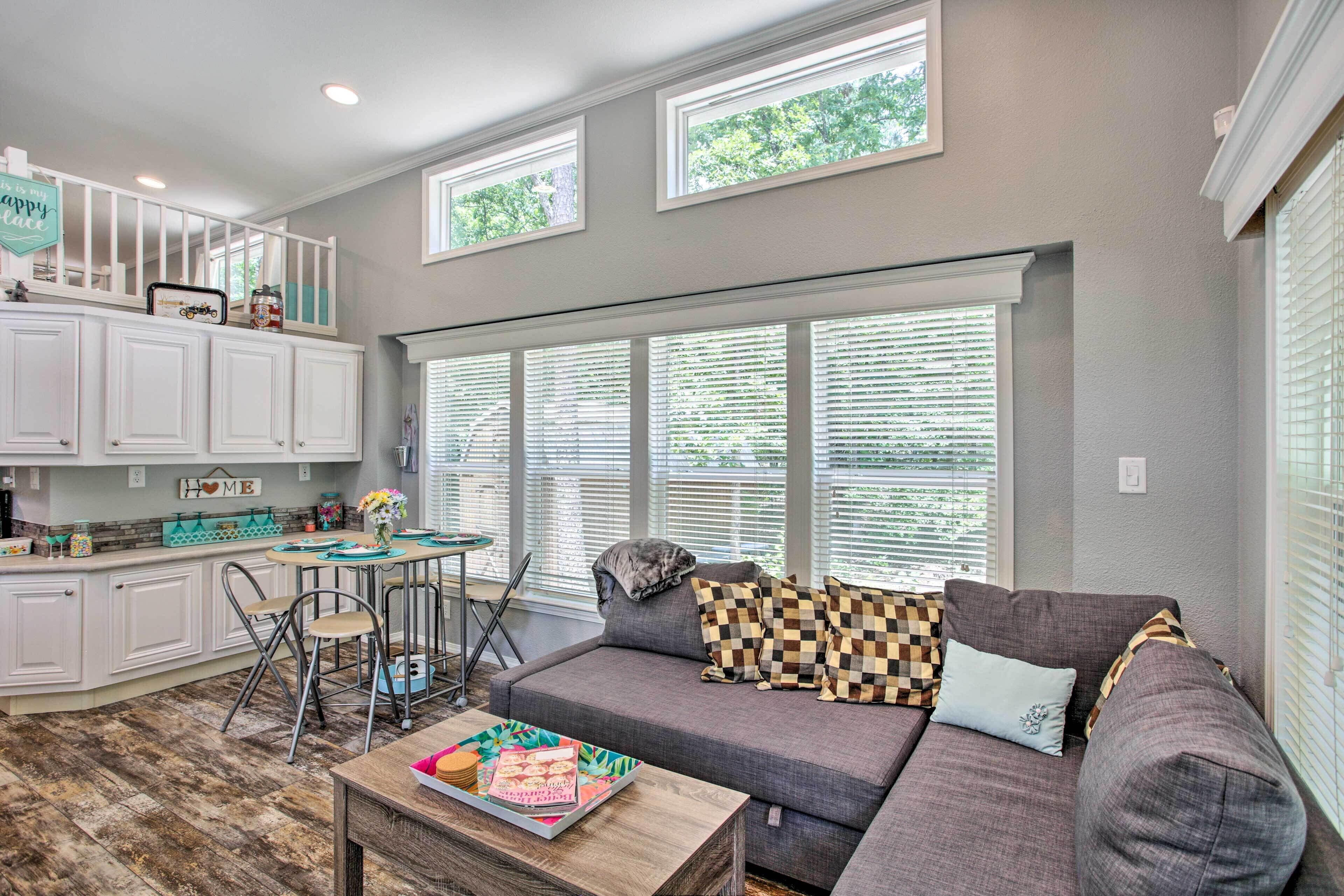 A spacious living room with a gray sofa, a coffee table, and a dining area with a small table and chairs. There are large windows with blinds, a kitchen area with white cabinets, and a staircase leading to an upper level.