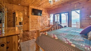 A wooden cabin bedroom with a bed, a ceiling fan, a TV mounted on the wall, and a view of the outside through large windows.