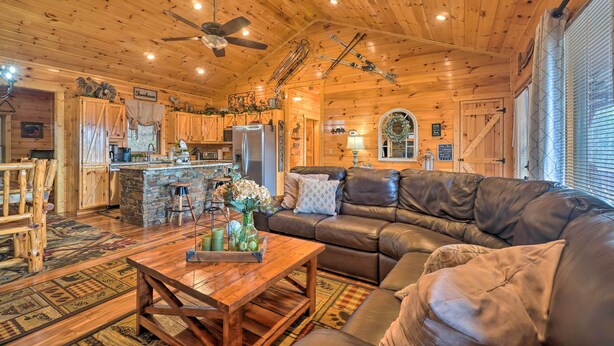 A cozy cabin interior with a wooden ceiling, a stone fireplace, a leather sofa, and a dining area with wooden furniture.