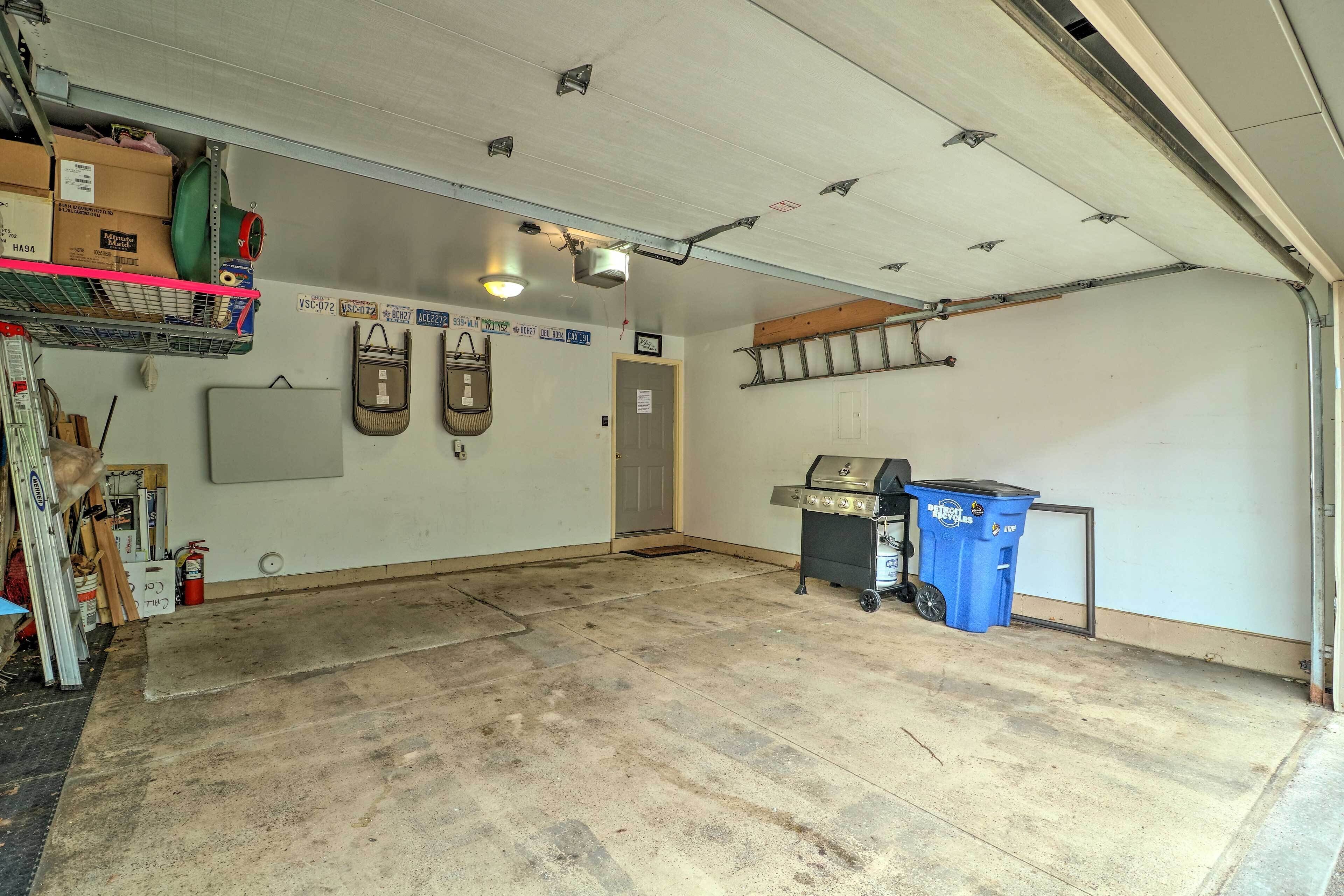Eine Garage mit Betonboden, Deckenventilator, an der Wand befestigtem Regal mit Kartons, einem blauen Mülleimer und einem Grill.