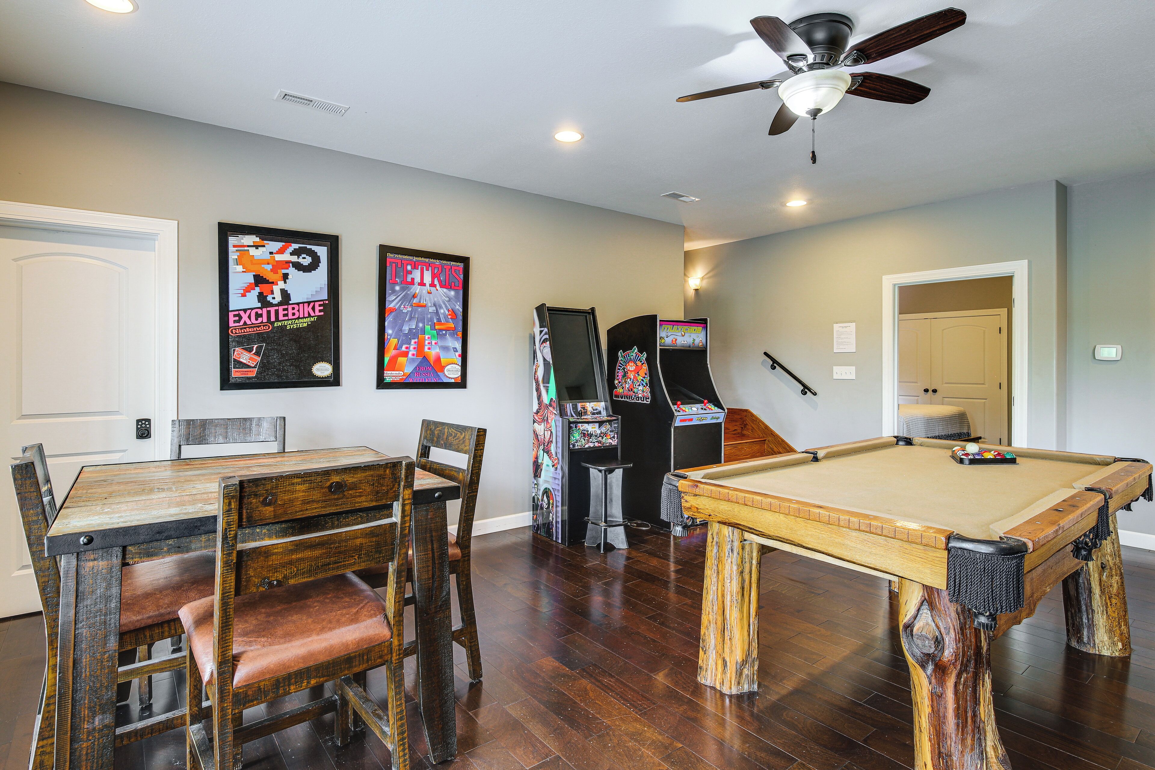 A room with a pool table, wooden chairs, a ceiling fan, arcade games, and framed posters on the wall.
