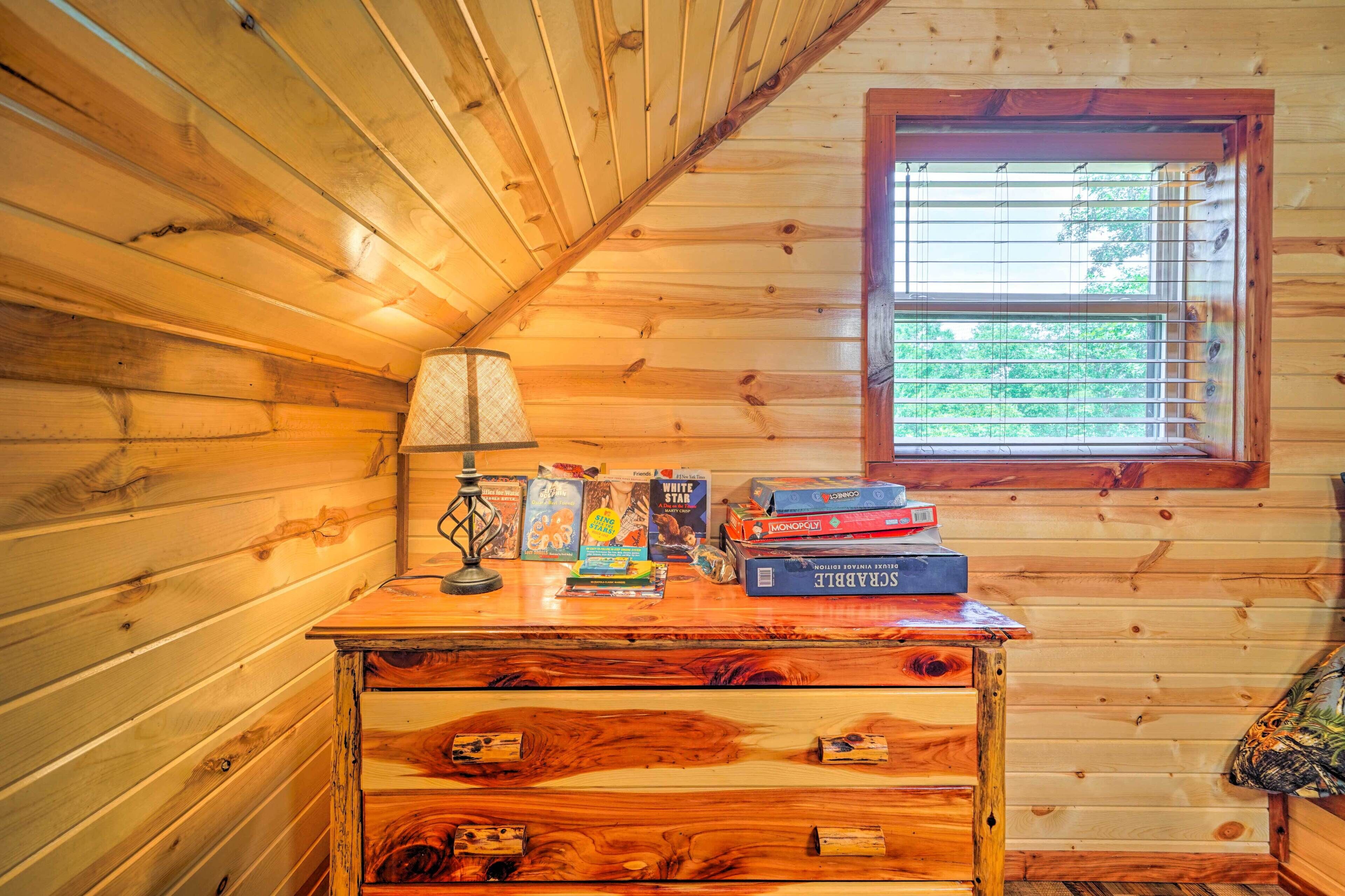 Une pièce de cabane en bois avec une fenêtre, un bureau avec des livres et une lampe, et un lit recouvert d’une couverture à motifs camouflage.