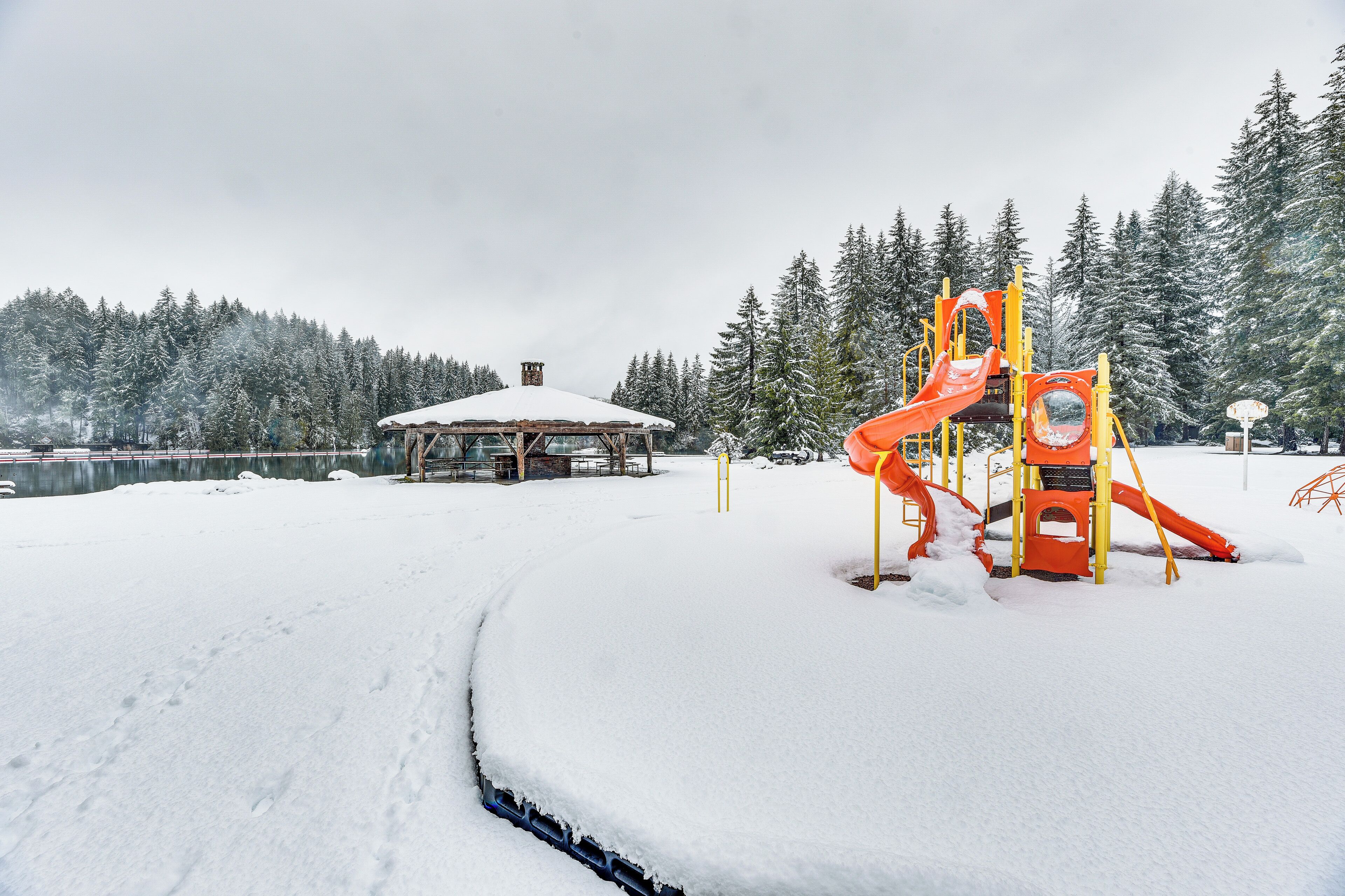 A snow-covered playground with a slide and climbing structures, surrounded by a forest and a building with a snow-covered roof.