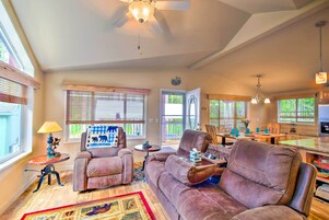 A spacious living room with a large sofa, a coffee table, and a fireplace. There is a dining area with a table and chairs, and a kitchen in the background.