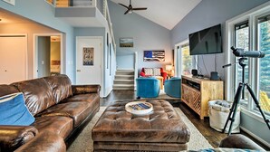 A spacious living room with a brown leather couch, a tufted ottoman, a blue armchair, and a wooden cabinet. A ceiling fan is visible, and a telescope is placed near a large window. - Fire Pit, Smart TV: Waterfront Charlevoix Home (Charlevoix)