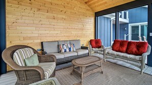A wooden-paneled room with wicker furniture, including a sofa and chairs, and a large window offering a view of a building outside. - Fire Pit, Smart TV: Waterfront Charlevoix Home (Charlevoix)
