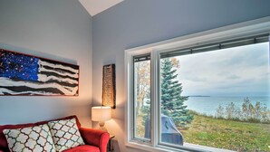 A living room with a red couch, a blue armchair, a wooden coffee table, a painting, and a view of a lake and trees. - Fire Pit, Smart TV: Waterfront Charlevoix Home (Charlevoix)