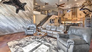 A spacious living area with leather furniture, a staircase, and a kitchen in the background. - Kansas Hunting Lodge: Pets & Large Groups Welcome! (Great Bend)