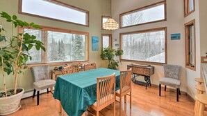 A dining area with a table set for six, surrounded by wooden chairs, large windows, and a hanging light fixture.