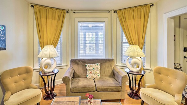 Un salon avec un canapé beige, deux fauteuils et une table basse agrémentée d’objets décoratifs. Deux lampes sont posées sur des tables d’appoint, et la pièce est dotée d’une fenêtre avec des rideaux.