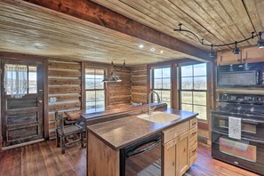 A rustic kitchen with wooden walls, a large island with a sink, a stove, and a view of the outdoors.