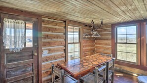 A rustic log cabin interior with a wooden dining table and chairs, large windows, and a chandelier. - Rustic & Rural Cabin in Dupuyer on Open 14 Acres! (Dupuyer)