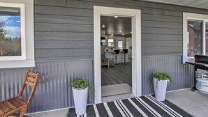 Fachada moderna con un muro gris, una silla de madera, dos plantas en macetas y un tapete a rayas blanco y negro que lleva a una puerta.