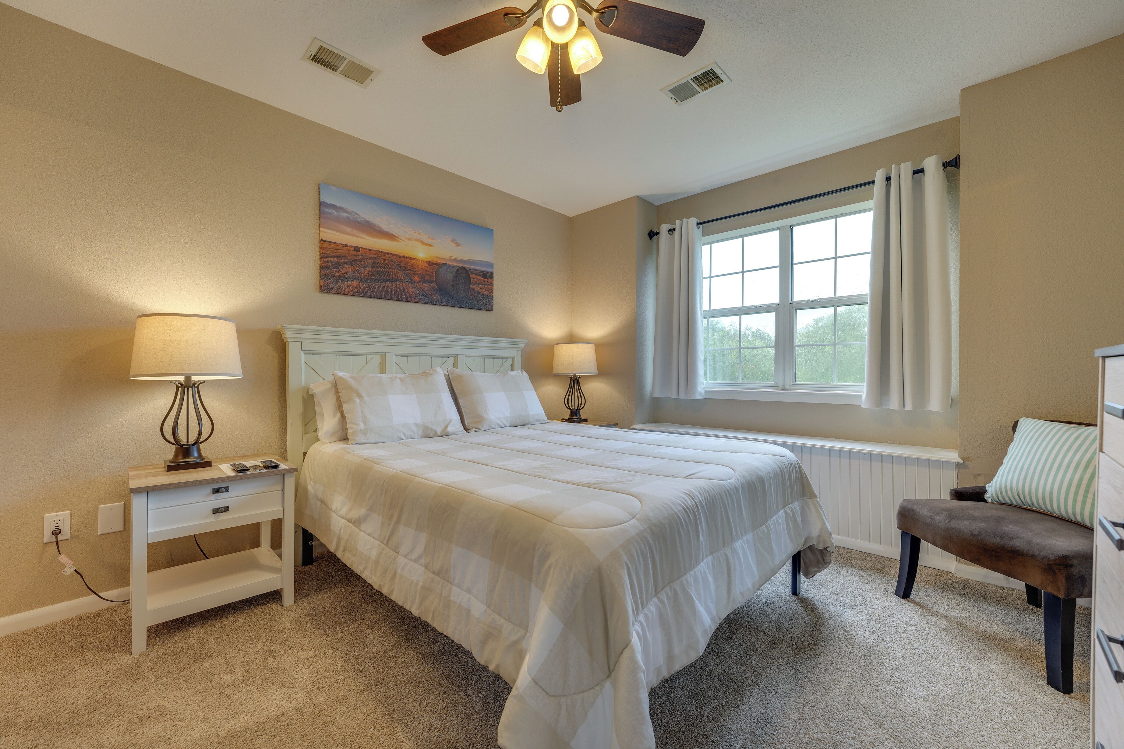 A bedroom with a bed, bedside lamps, a ceiling fan, a window with curtains, and a painting on the wall.