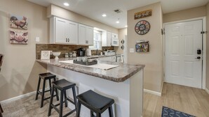 A kitchen with white cabinets, a granite countertop, and a bar area with stools. There are wall decorations, including a rooster painting and a clock.