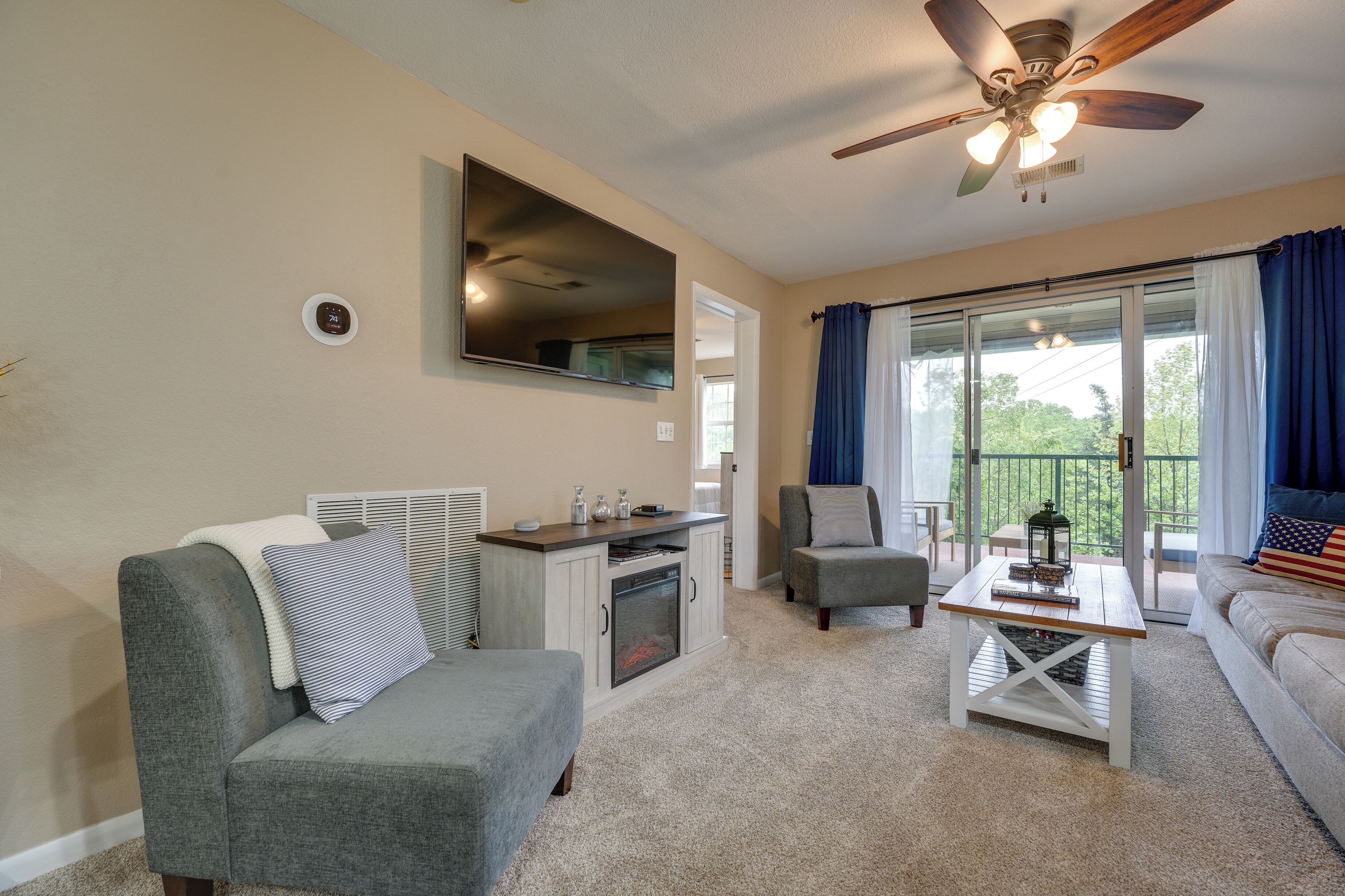 A living room with a gray sofa, a coffee table, a gray armchair, a fireplace, a ceiling fan, and a large window with blue curtains.