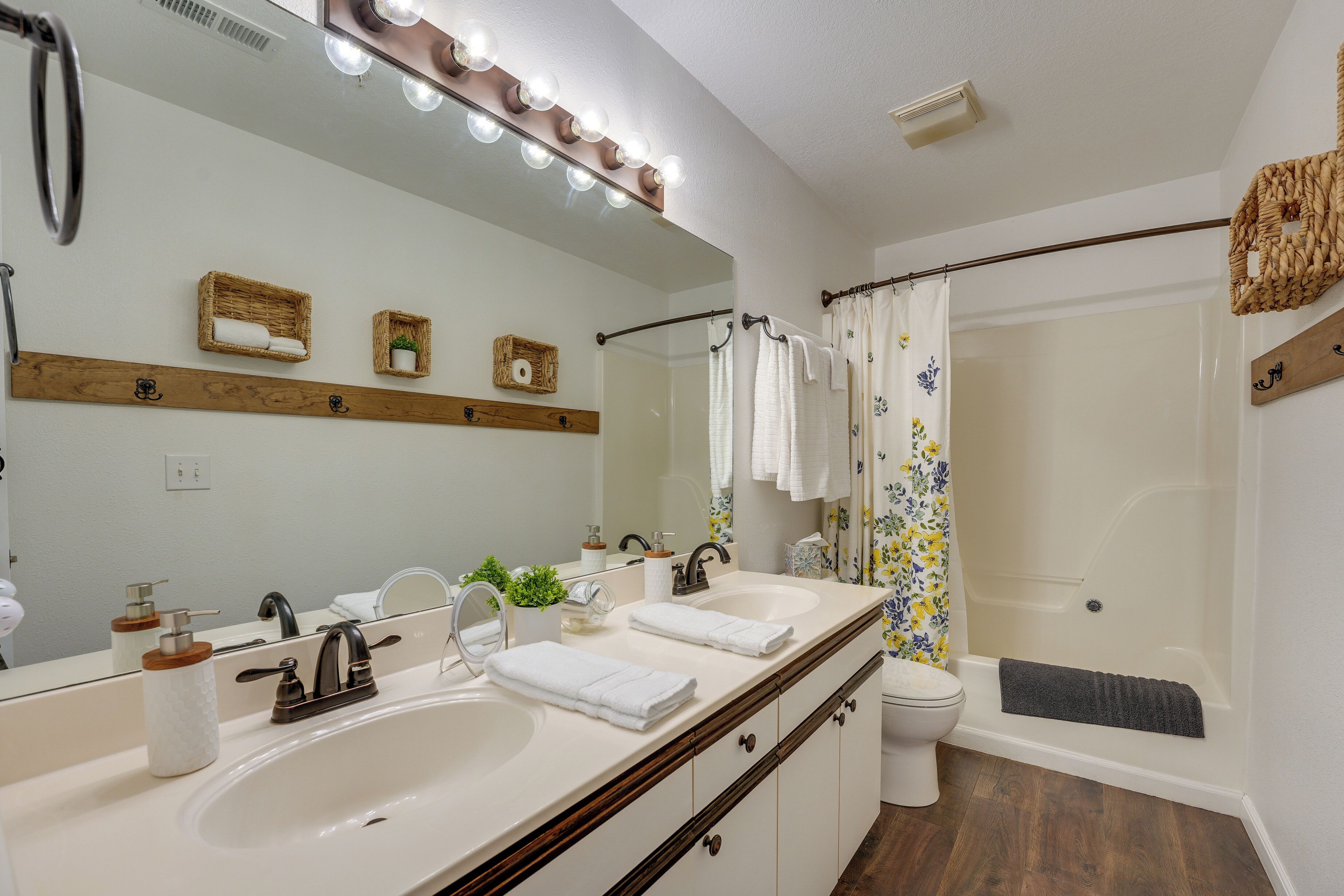 A bathroom with two sinks, a mirror with lights, a shower with a floral curtain, and wooden shelves with towels and a plant.