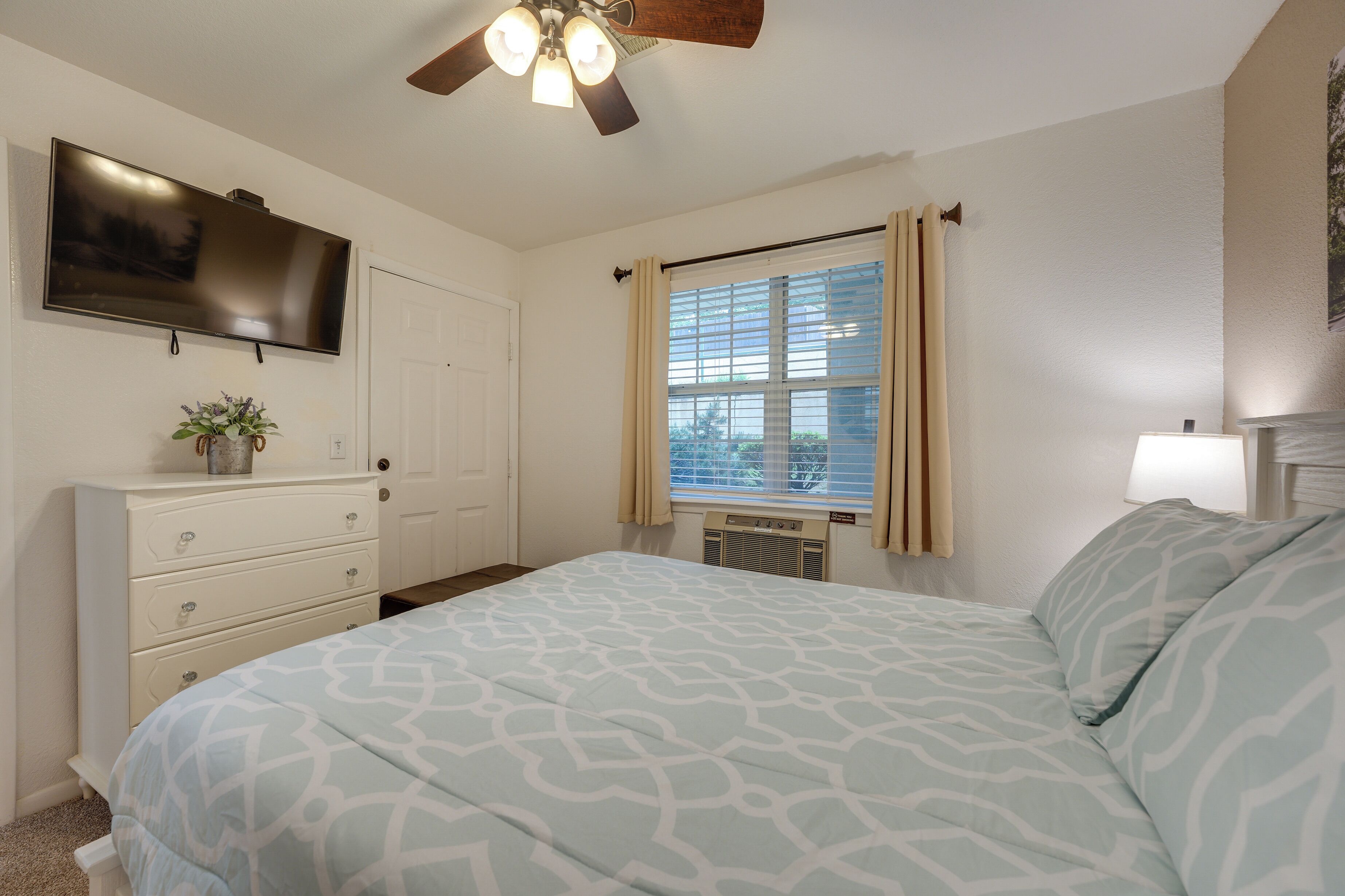 Une chambre à coucher comprenant un lit, un ventilateur de plafond, une télévision, une commode, une fenêtre avec des rideaux et un radiateur.