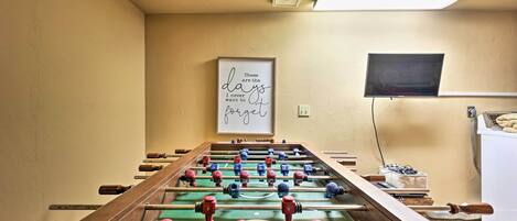 A foosball table in a room with beige walls, a framed inspirational quote, and a television mounted on the wall.