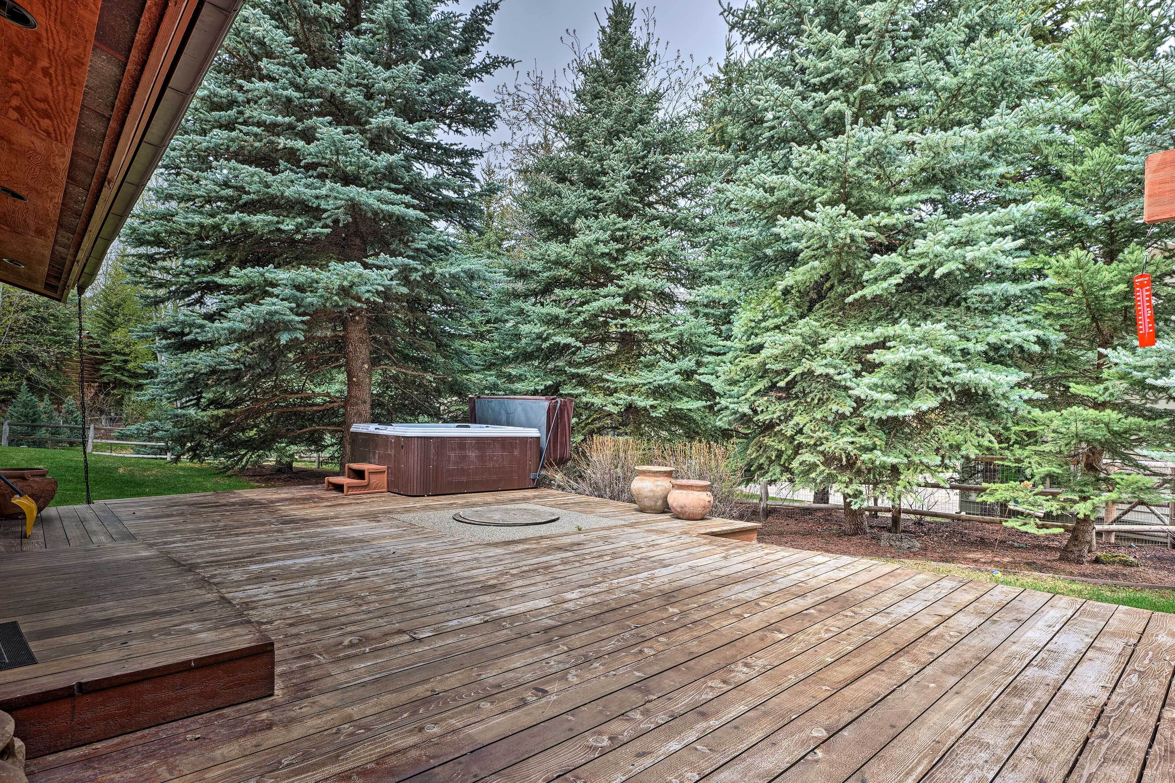 Une terrasse en bois avec un jacuzzi et un espace salon, entourée de grands arbres à feuilles persistantes.
