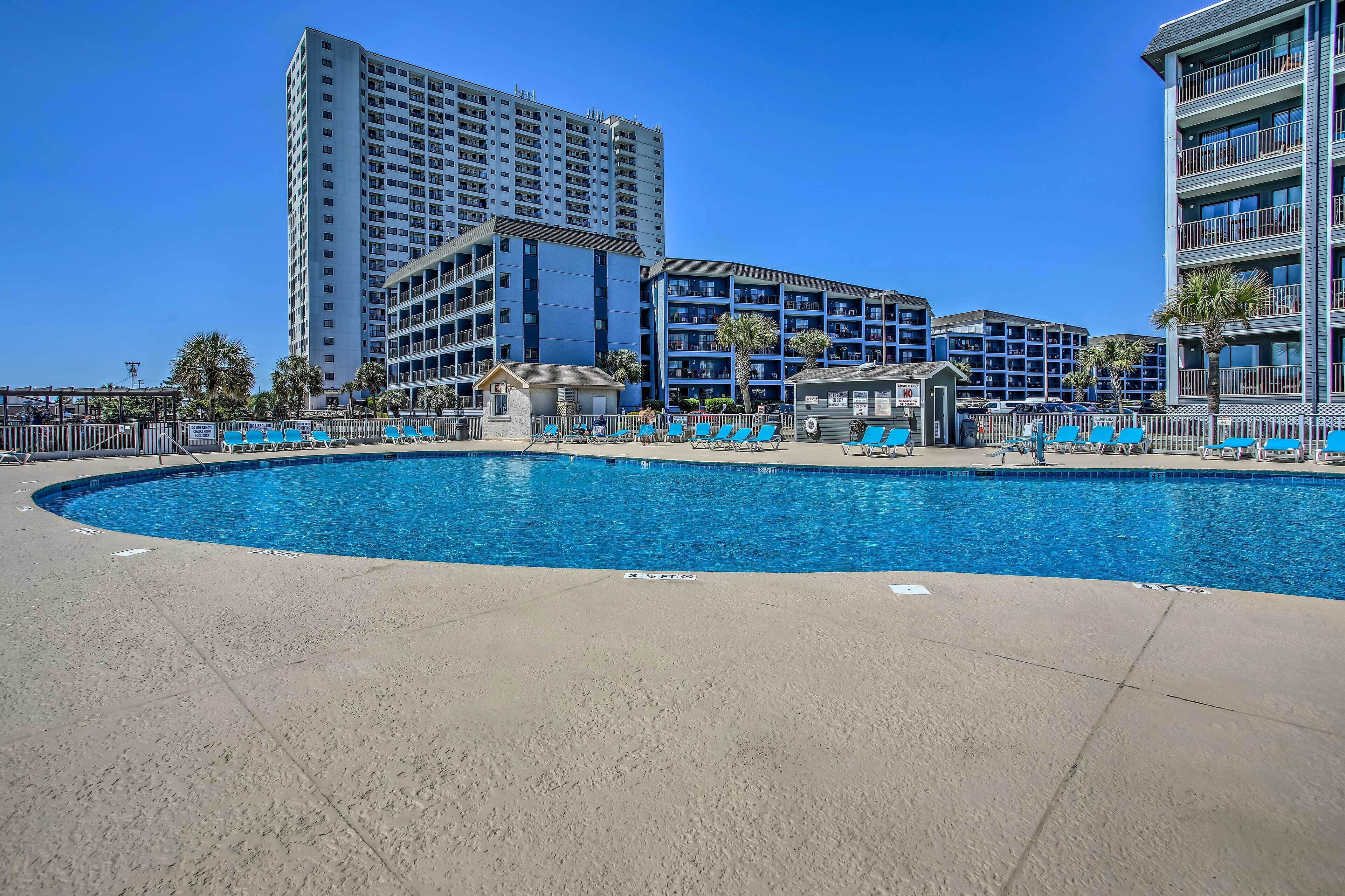 A hotel with a large swimming pool surrounded by lounge chairs and palm trees.